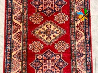 Azerbaijan Tappeti Milano Un tappeto a motivi tradizionali con disegni geometrici nei toni del rosso, blu e beige, esposto su un pavimento piastrellato con un motivo a farfalla nell'angolo in alto a destra.