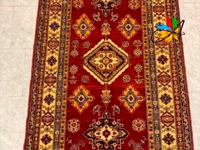 Azerbaijan Tappeti Milano Un tappeto decorativo con motivi intricati e un design tradizionale su un pavimento piastrellato, con un motivo a farfalla colorato in un angolo.