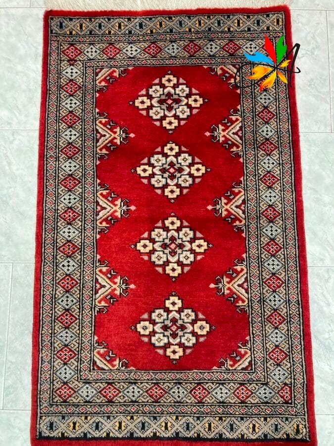 Azerbaijan Tappeti Milano Un tradizionale tappeto rosso con intricati motivi geometrici e dettagli sui bordi visualizzati su un pavimento di colore chiaro, accompagnato da un'illustrazione digitale di una farfalla colorata nell'angolo in alto a destra per un tocco artistico.