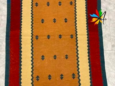 Azerbaijan Tappeti Milano Un tappeto tradizionale con un campo centrale marrone chiaro, motivi a rombi e un bordo geometrico multicolore disposto su un pavimento piastrellato, con una piccola farfalla artificiale colorata posizionata nell'angolo in alto a destra.