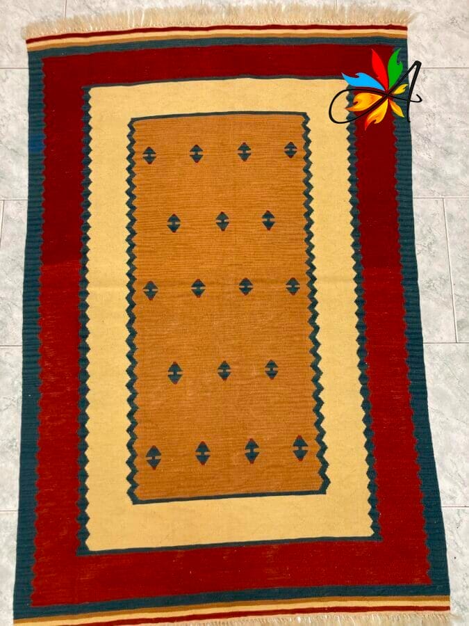Azerbaijan Tappeti Milano Un tappeto tradizionale con un campo centrale marrone chiaro, motivi a rombi e un bordo geometrico multicolore disposto su un pavimento piastrellato, con una piccola farfalla artificiale colorata posizionata nell'angolo in alto a destra.
