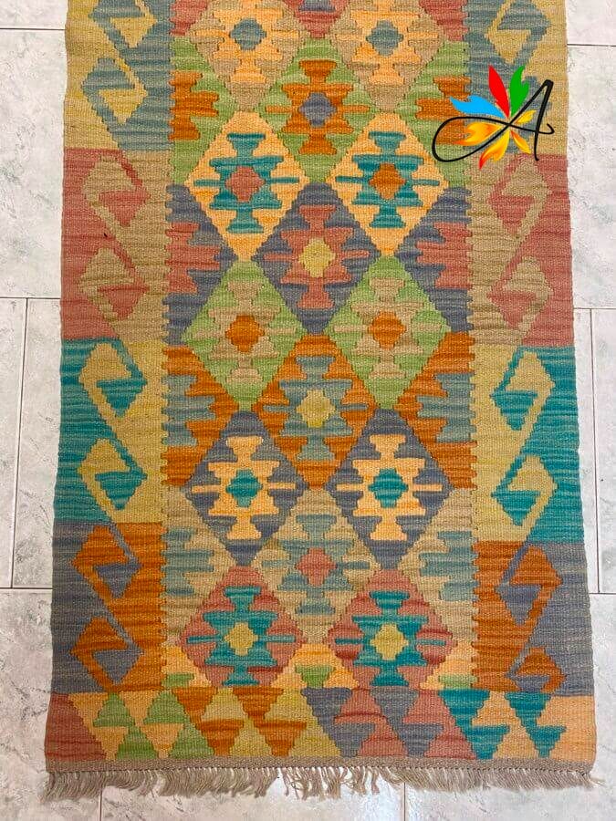 Azerbaijan Tappeti Milano Tappeto colorato con motivi geometrici a rombi e triangoli nei toni del verde, blu, arancio, giallo e rosso. Su un'estremità sono visibili delle frange e nell'angolo in alto a destra compare un logo multicolore.