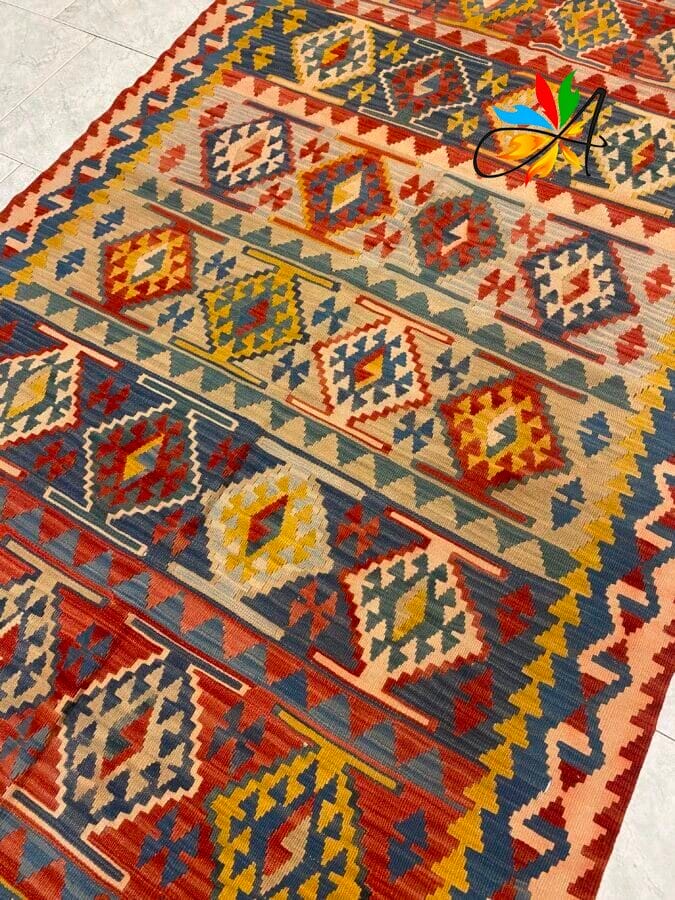 Azerbaijan Tappeti Milano Tappeto a motivi geometrici e colorati, con disegni a rombi e a zig zag nei toni del rosso, giallo, blu, verde e beige, steso su un pavimento piastrellato chiaro. Nell'angolo in alto a destra è presente il logo di una farfalla colorata.