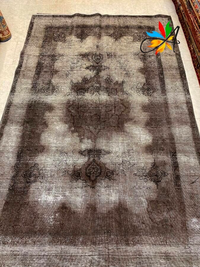 Azerbaijan Tappeti Milano Un grande tappeto rettangolare sbiadito con un motivo floreale centrale in tonalità di grigio e nero. Un logo a forma di foglia colorata si trova nell'angolo in alto a destra.