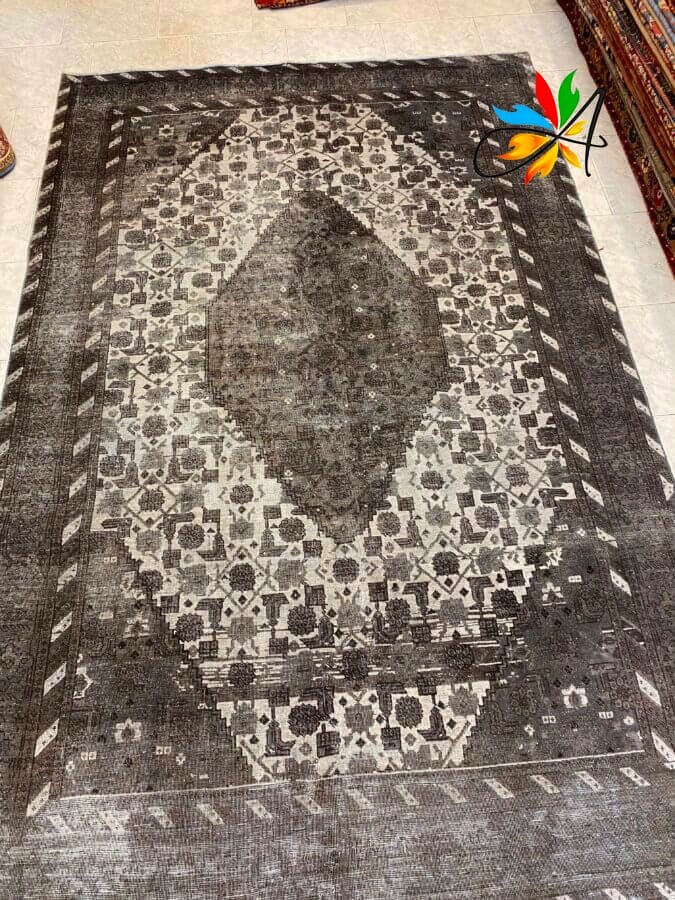 Azerbaijan Tappeti Milano Un tappeto grigio e nero con motivo geometrico e forma centrale a diamante, esposto su un pavimento chiaro. Un logo colorato è nell'angolo in alto a destra.