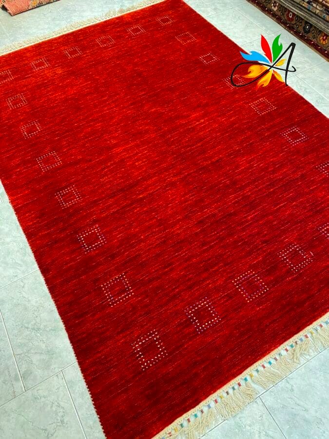 Azerbaijan Tappeti Milano Un tappeto rosso con un sottile motivo quadrato su un pavimento chiaro. Un disegno multicolore è visibile nell'angolo in alto a destra.