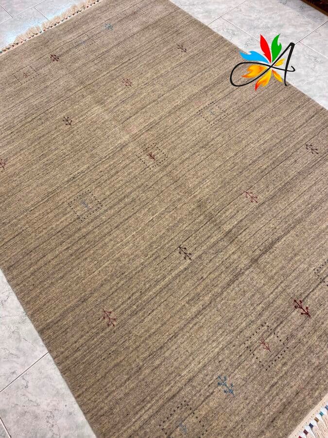 Azerbaijan Tappeti Milano Un tappeto beige con un motivo strutturato caratterizzato da piccoli disegni sparsi e un logo colorato nell'angolo in alto a destra, posizionato su un pavimento piastrellato.