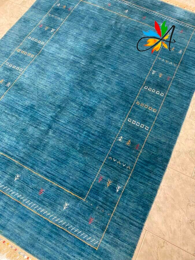 Azerbaijan Tappeti Milano Tappeto blu con un motivo di bordo sottile con piccoli motivi geometrici e naturali. Un logo colorato con la lettera "A" è nell'angolo in alto a destra. Tappeto posizionato su un pavimento piastrellato chiaro.