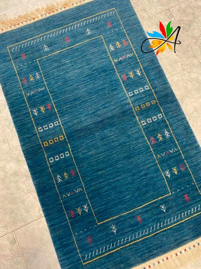 Azerbaijan Tappeti Milano Tappeto rettangolare blu con motivi geometrici e astratti in rosso, giallo e bianco, bordato da frange. Posizionato su un pavimento piastrellato, con un logo colorato nell'angolo in alto a destra.
