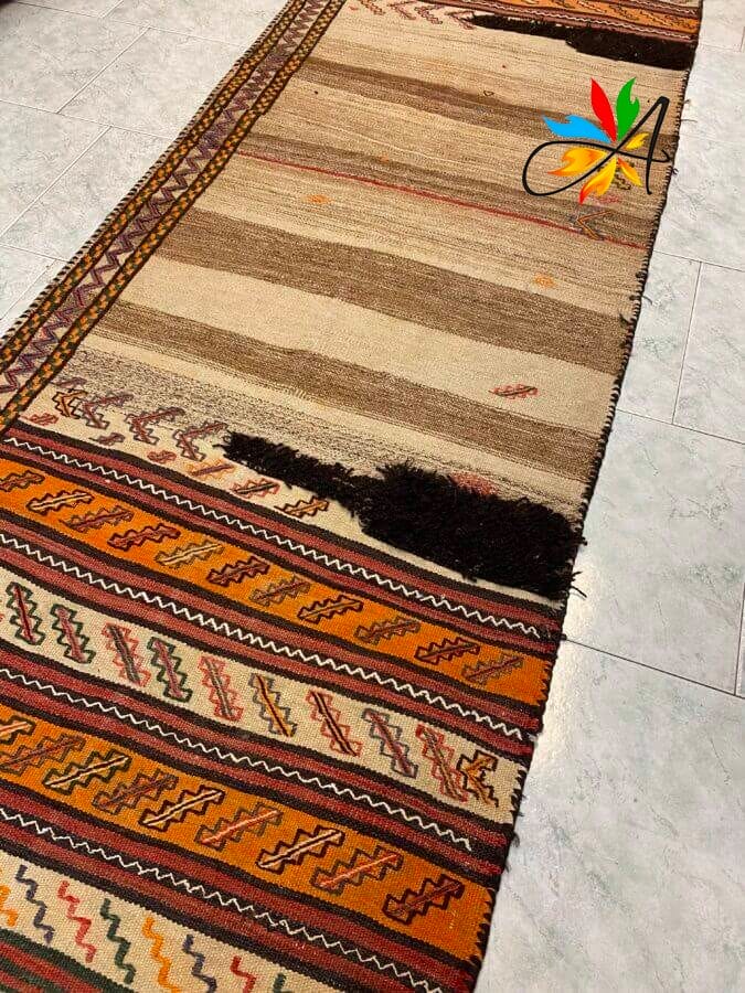 Azerbaijan Tappeti Milano Un lungo tappeto a fantasia con toni terrosi e disegni tribali è steso su un pavimento piastrellato. Un elemento decorativo colorato è su un lato.