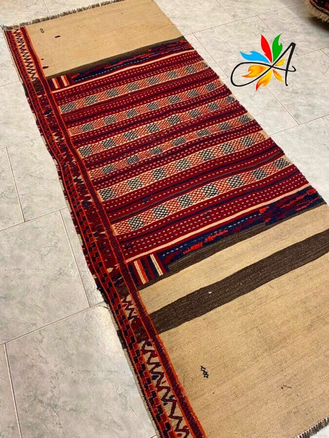 Azerbaijan Tappeti Milano Un tappeto rettangolare intrecciato con motivi geometrici rossi, beige e neri su un pavimento piastrellato. Un logo colorato è nell'angolo in alto a destra.