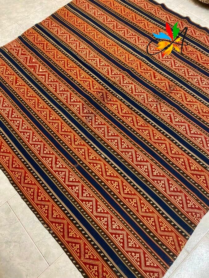 Azerbaijan Tappeti Milano Un tessuto intrecciato con motivi geometrici ripetuti a strisce rosse, marroni e nere. Un logo a forma di foglia colorata si trova nell'angolo in alto a destra.