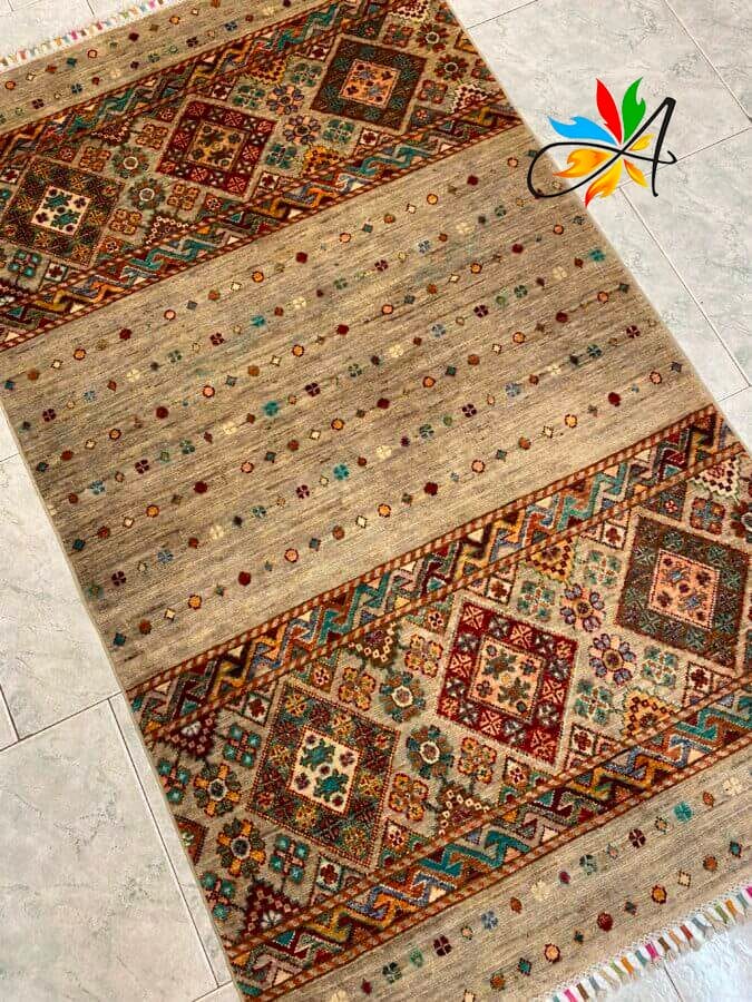 Azerbaijan Tappeti Milano Un tappeto con motivi geometrici e floreali in rosso, blu e verde su uno sfondo beige, esposto su un pavimento piastrellato. Un logo a forma di foglia colorata con la lettera "A" è nell'angolo in alto a destra.