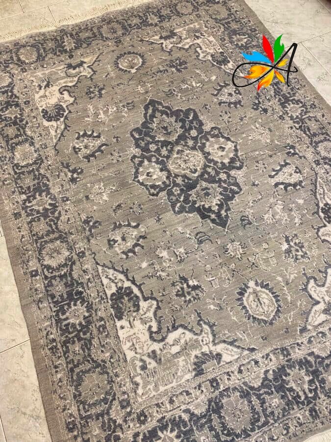 Azerbaijan Tappeti Milano Un tappeto rettangolare con un mix di toni di grigio e blu scuro, decorato con motivi floreali e geometrici, è adagiato su un pavimento piastrellato.