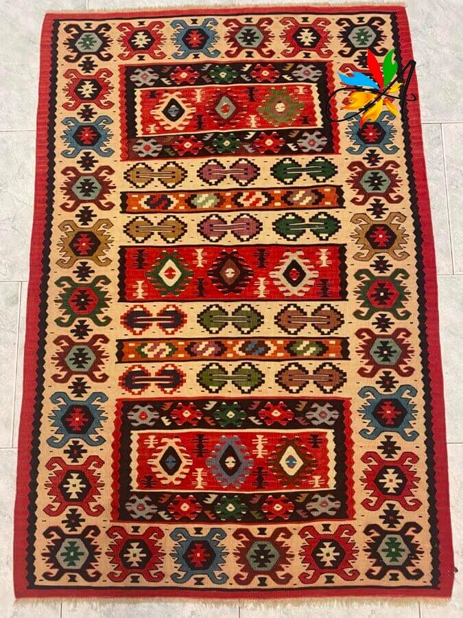 Azerbaijan Tappeti Milano Tappeto con motivi geometrici colorati, rosso, beige, verde e blu su un pavimento piastrellato chiaro.