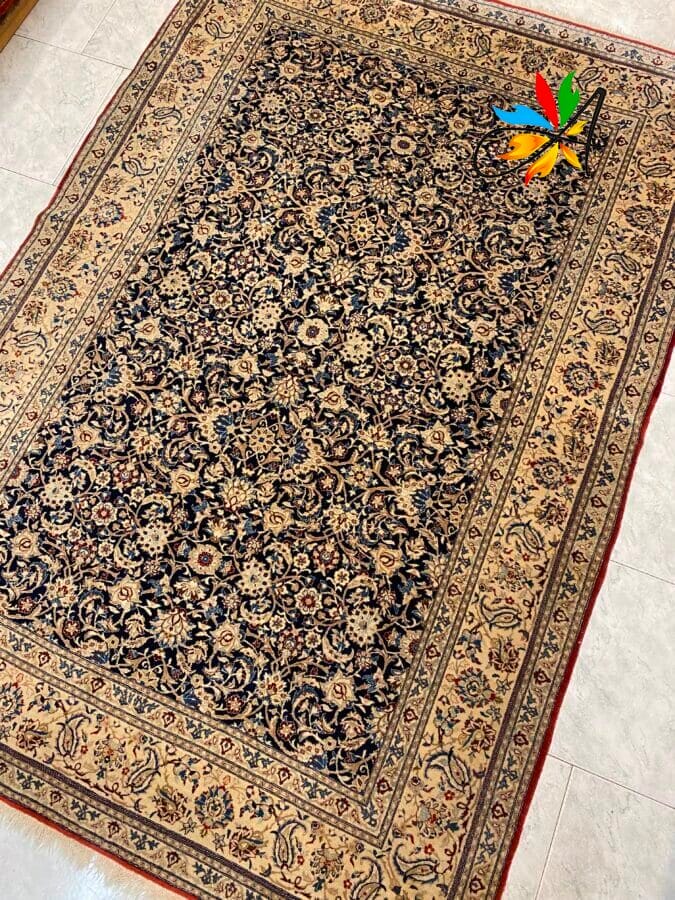 Azerbaijan Tappeti Milano Un tappeto rettangolare, decorato in stile persiano con intricati motivi floreali in toni scuri e chiari, bordato da un bordo decorativo, su un pavimento in piastrelle chiare. Un logo colorato è nell'angolo in alto a destra.