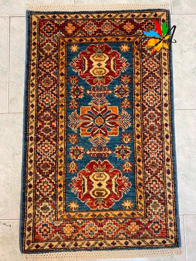 Azerbaijan Tappeti Milano Un tappeto orientale rettangolare con centro blu, motivi geometrici rossi e dorati e bordo decorato con motivi beige.