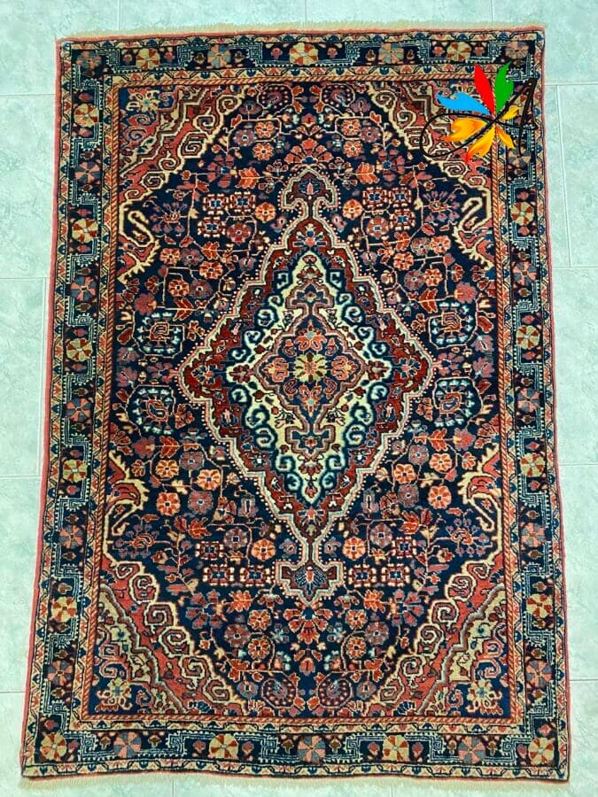 Azerbaijan Tappeti Milano Tappeto persiano decorato con intricati motivi floreali nei toni del rosso, blu e crema, adagiato su un pavimento piastrellato chiaro.