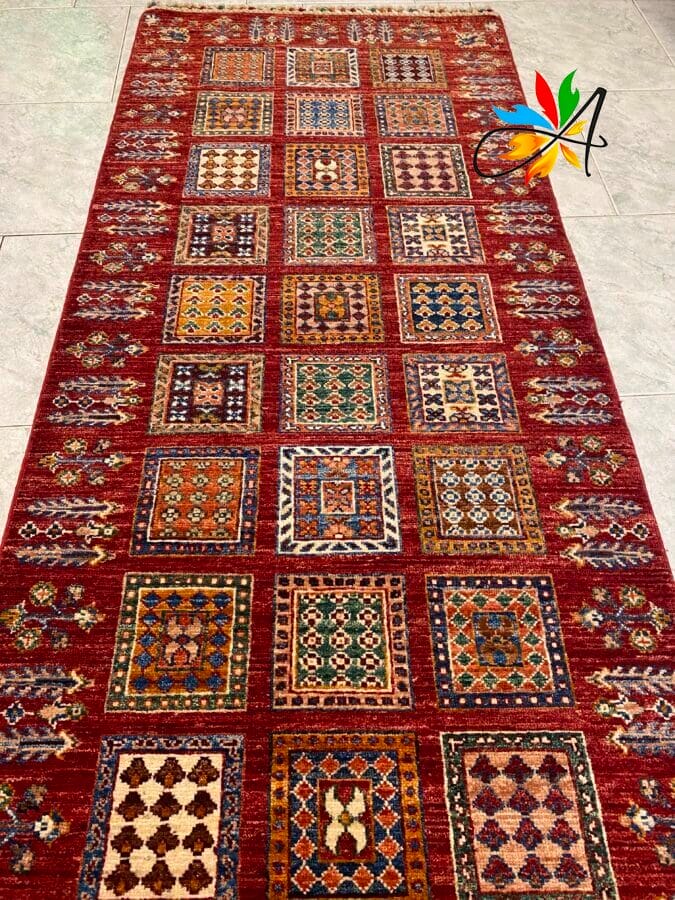 Azerbaijan Tappeti Milano Un tappeto rettangolare con colorati motivi geometrici quadrati su sfondo rosso, adagiato su un pavimento piastrellato.