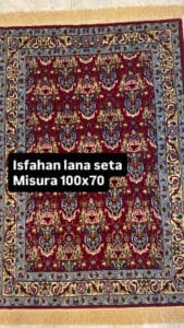 Azerbaijan Tappeti Milano Tappeto rettangolare in stile persiano con un ricco sfondo rosso, motivi floreali ornamentali e un dettagliato bordo blu e beige. Il testo in sovrimpressione recita: "Isfahan lana seta Misura 100x70.