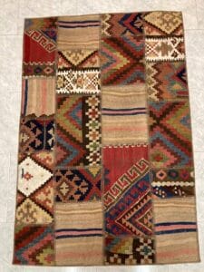 Azerbaijan Tappeti Milano Un tappeto patchwork multicolore con vari motivi geometrici disposti su un pavimento piastrellato, caratterizzato da una piccola pila di oggetti colorati nell'angolo in alto a destra.