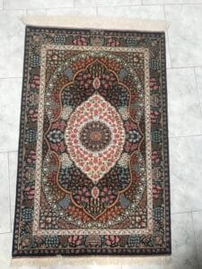 Azerbaijan Tappeti Milano Un tradizionale tappeto decorato esposto su un pavimento piastrellato con un disegno a forma di foglia nell'angolo che si sovrappone all'immagine.