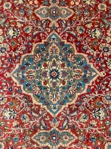 Azerbaijan Tappeti Milano Un tappeto persiano riccamente dettagliato caratterizzato da intricati motivi floreali e geometrici, con una vibrante tavolozza di rosso, blu e crema e una piccola farfalla artificiale appoggiata sulla sua superficie.