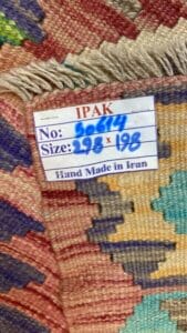 Azerbaijan Tappeti Milano Primo piano di un tappeto tessuto con un'etichetta che recita: "IPAK, No: 50644, Dimensioni: 298 x 198, Fatto a mano in Iran". Il tappeto colorato presenta motivi geometrici e un bordo con frange.