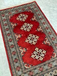 Azerbaijan Tappeti Milano Un tappeto tradizionale rosso e beige di ottima fattura con intricati motivi geometrici e bordi, posato su un pavimento piastrellato chiaro, con un logo a forma di fiore nell'angolo che suggerisce un marchio o il marchio di un negozio.