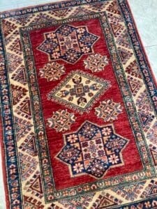 Azerbaijan Tappeti Milano Un tappeto a motivi tradizionali con disegni geometrici nei colori rosso, blu e crema, esposto su un pavimento.
