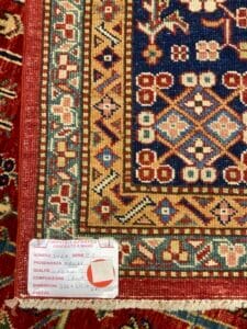 Azerbaijan Tappeti Milano Una sezione dettagliata di un tappeto dai motivi vivaci che mostra disegni intricati e un'etichetta allegata che ne indica l'autenticità, l'origine, la qualità, le dimensioni e il prezzo.