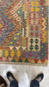 Azerbaijan Tappeti Milano Tappeto a motivi colorati con forme geometriche in giallo, blu, arancione, rosa e verde. Parte delle gambe e delle scarpe di una persona sono visibili in piedi su un pavimento piastrellato bianco nella parte inferiore dell'immagine.