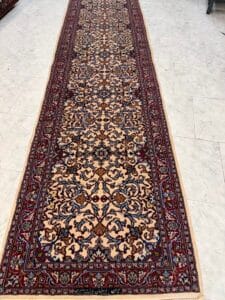 Azerbaijan Tappeti Milano Un lungo tappeto persiano decorato con intricati motivi floreali e di viti nei toni del rosso, del blu e del beige, posto su un pavimento piastrellato chiaro. Un logo colorato e stilizzato appare nell'angolo in alto a destra.