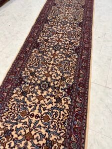 Azerbaijan Tappeti Milano Un tappeto lungo e decorato con un intricato motivo floreale nei toni del beige, del rosso e del blu giace su un pavimento piastrellato chiaro. Un logo colorato a forma di foglia con il numero 74 appare nell'angolo in alto a destra.