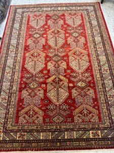 Azerbaijan Tappeti Milano Tappeto persiano tradizionale rosso con intricati motivi geometrici e floreali nei toni del crema, del giallo e del nero. Il logo della foglia multicolore e la scritta in nero compaiono nell'angolo in alto a destra.