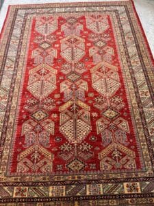 Azerbaijan Tappeti Milano Tappeto tradizionale a fantasia rossa con disegni geometrici in crema, giallo e blu, bordati da motivi intricati. Nell'angolo in alto a destra compare il logo di una foglia e di una penna colorate.