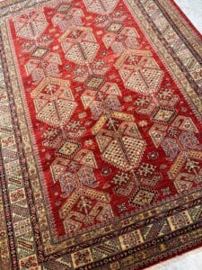 Azerbaijan Tappeti Milano Tappeto a fantasia rossa con intricati disegni geometrici nei toni del crema, del blu e del giallo, delimitati da motivi dettagliati; nell'angolo in alto a destra un logo colorato con una farfalla multicolore.