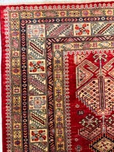 Azerbaijan Tappeti Milano Primo piano di un tappeto tradizionale con intricati motivi geometrici in rosso, crema e blu scuro. I bordi presentano motivi dettagliati e l'angolo mostra un piccolo pappagallo ricamato che tiene in mano un peperoncino.