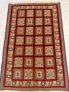 Azerbaijan Tappeti Milano Un tappeto rettangolare con sfondo rosso e un motivo a griglia di motivi geometrici beige e blu, delimitato da disegni intricati in crema, rosso e blu. Nell'angolo superiore destro compare il logo di una farfalla colorata.