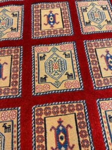 Azerbaijan Tappeti Milano Un tappeto a fantasia rossa presenta disegni rettangolari con forme geometriche e motivi di piccoli animali, bordati di blu e beige, con un logo colorato nell'angolo in alto a destra.