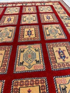 Azerbaijan Tappeti Milano Primo piano dettagliato di un tappeto rosso con motivi geometrici in beige, blu e nero. Il tappeto presenta quadrati bordati con motivi intricati. Un logo colorato è visibile nell'angolo in alto a destra.