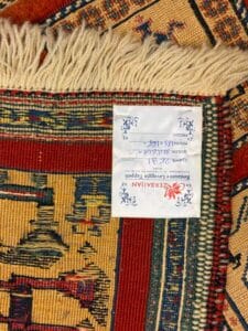 Azerbaijan Tappeti Milano Primo piano dell'angolo di un tappeto a fantasia che mostra le frange, la tessitura dettagliata e un'etichetta bianca con testo blu e rosso, parzialmente in lingua straniera, attaccata sul retro. Un logo colorato si trova nell'angolo in alto a destra.