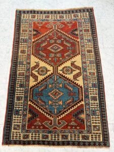Azerbaijan Tappeti Milano Tappeto rettangolare con motivi geometrici nei toni del rosso, del blu, del beige e del marrone, caratterizzato da due grandi medaglioni esagonali e da intricati disegni di bordo, esposto su un pavimento piastrellato chiaro.