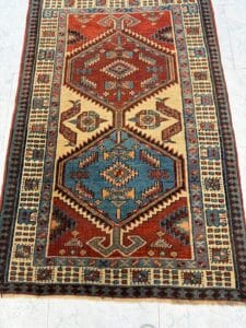Azerbaijan Tappeti Milano Tappeto rettangolare con motivi geometrici nei toni del ruggine, blu, crema e marrone, caratterizzato da due grandi esagoni al centro e da un bordo decorativo. Nell'angolo compare un logo colorato a forma di stella.
