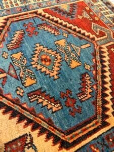 Azerbaijan Tappeti Milano Primo piano di un tappeto tradizionale con motivi geometrici in blu, rosso, crema e arancione. Nell'angolo in alto a destra compaiono una foglia colorata e il logo "A" stilizzato.