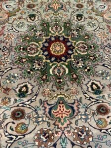 Azerbaijan Tappeti Milano Tappeto persiano dettagliato e ornato, con un intricato medaglione floreale centrale nei toni del verde, del blu e del rosso, circondato da motivi elaborati. Nell'angolo in alto a destra si trova un logo astratto colorato con una "A" nera.