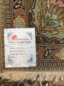 Azerbaijan Tappeti Milano Primo piano di un angolo di tappeto con un'etichetta allegata che recita "Azerbaijan Restauro e Lavaggio Tappeti" e dettagli scritti a mano su qualità, dimensioni e prezzo. Il tappeto presenta motivi intricati e bordi sfrangiati.