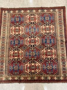 Azerbaijan Tappeti Milano Tappeto a motivi colorati con forme geometriche nei toni del rosso, del blu e del crema su un pavimento piastrellato.