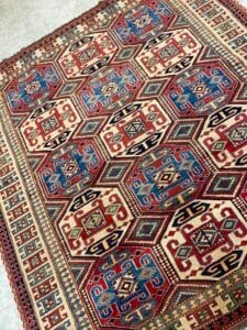 Azerbaijan Tappeti Milano Tappeto a motivi geometrici con colori rosso, blu e crema, oltre a un logo a forma di foglia colorata nell'angolo.