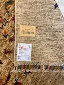 Azerbaijan Tappeti Milano Primo piano di un angolo di tappeto intrecciato beige con etichette e nappe colorate, posato su un tappeto a motivi.